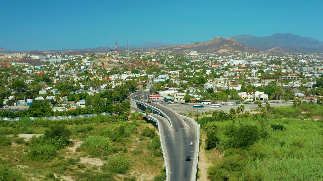 Wide San Jose Del Cabo view with the highway and bridge in the lower frame, green scrub and trees on both sides, white buildings across the town, mountains behind, and no visible people.