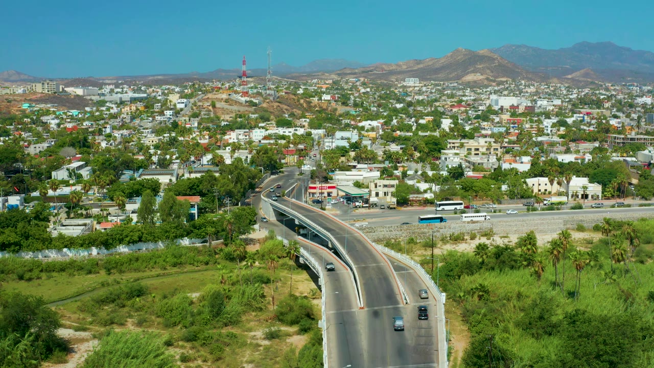 Aerial frame in San Jose Del Cabo with the curving overpass centered lower in frame, traffic on the main road below, hillside neighborhoods, distant mountains, and no visible people.