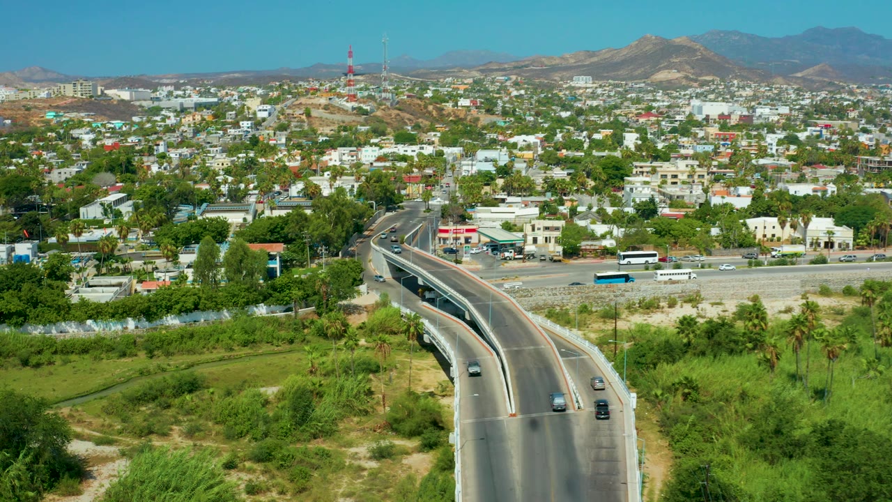 High aerial view of San Jose Del Cabo traffic on an elevated roadway, with a bus on the surface road, dense green trees, white buildings, dry ridges, and no visible people.