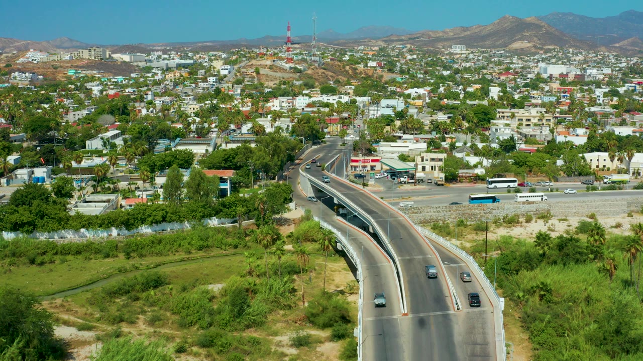 Wide aerial view of traffic, an overpass, green trees, buildings, and hills.