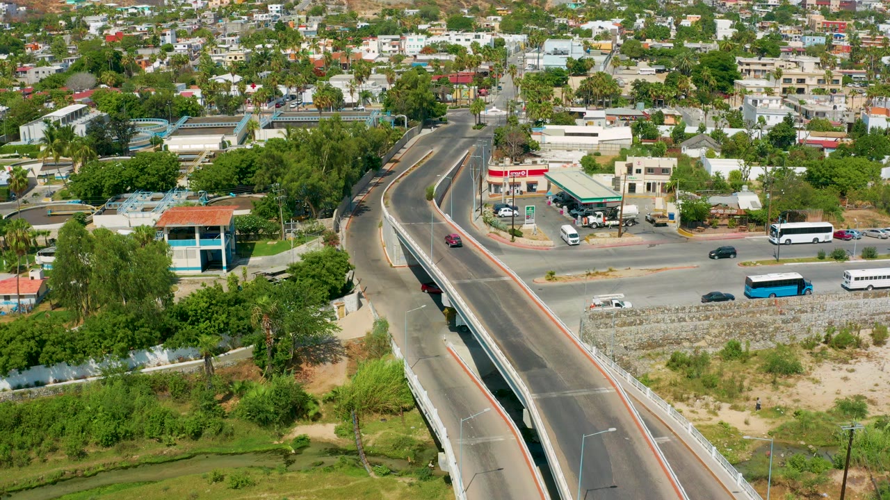 Aerial view in San Jose Del Cabo, Mexico with the bridge centered, a white bus near the roadside, a blue bus on the lower road, and neighborhood buildings in the background, with no visible people.