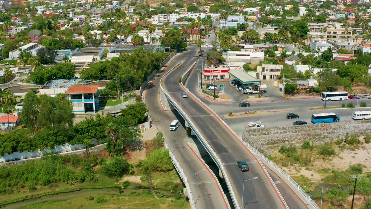 Aerial frame over San Jose Del Cabo showing a diagonal road overpass, moving vehicles, low buildings, residential blocks, green trees, scrub vegetation, and no visible people.