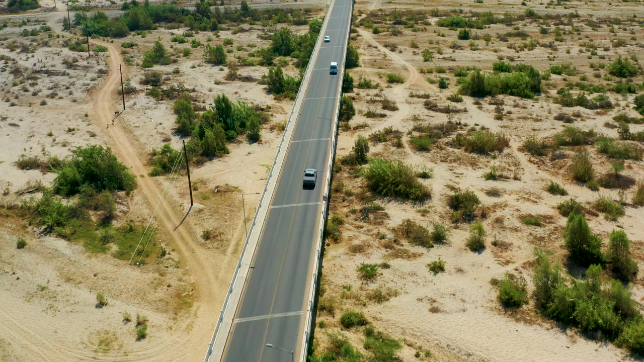 Aerial frame from San Jose del Cabo, Mexico, showing a dark vehicle traveling on the highway beside a branching dirt road, scattered shrubs, and denser green bushes, with no visible people.