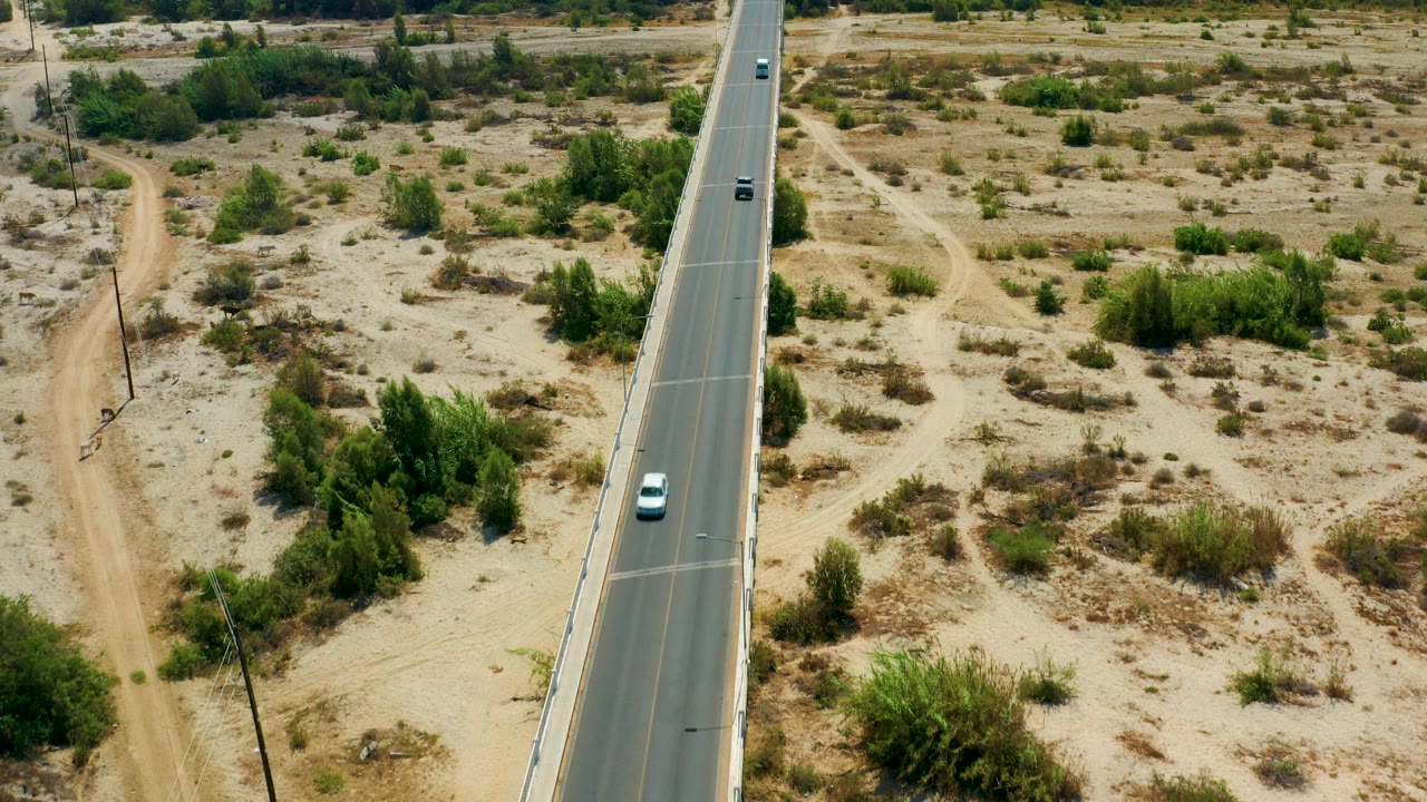 Aerial view in San Jose Del Cabo, Mexico, showing a paved road through sandy scrubland, scattered green foliage, dirt tracks, utility poles, and no visible people.
