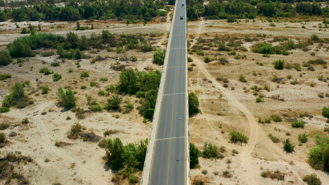 The overhead angle shows the highway in San Jose Del Cabo with faint dirt paths cutting through dry ground, denser green shrubs near the road edge, and no visible people.