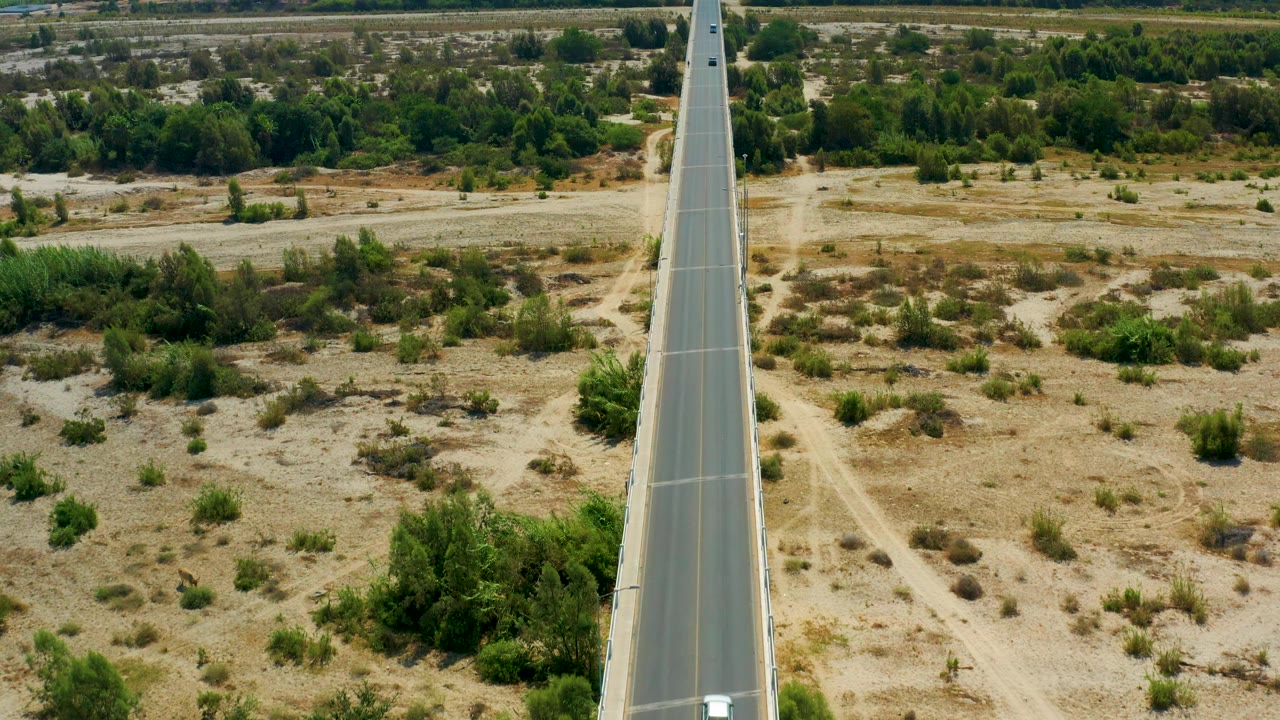 High view of a straight highway in San Jose Del Cabo, Mexico, with a small car visible near the upper part of the road and dry scrubland spreading to both sides, no visible people.