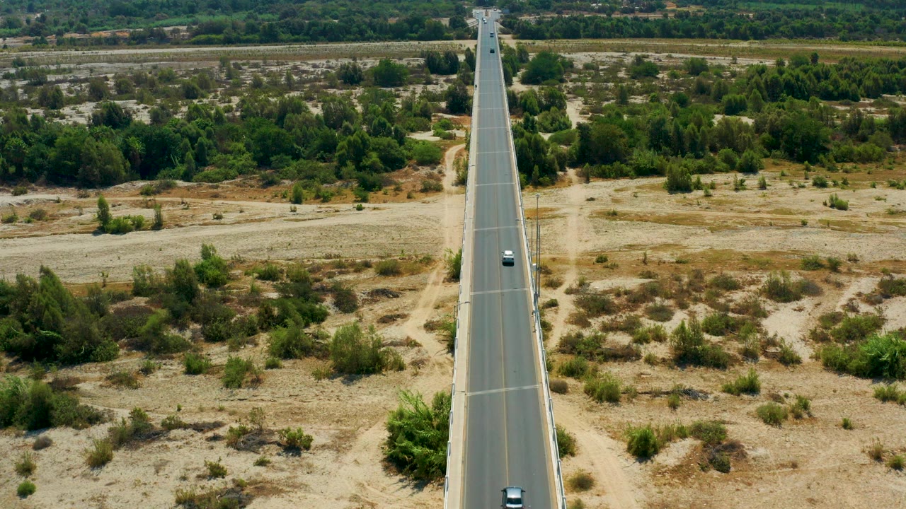 Opening aerial-style frame in San Jose Del Cabo, Mexico showing a straight road centered between dry fields, green brush, distant trees, and no visible people.