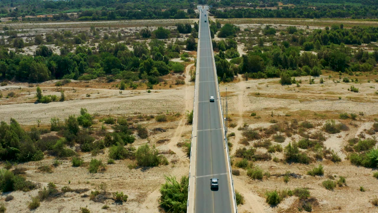 This San Jose Del Cabo frame keeps the long road corridor in view, with a dark vehicle on the lane, pale open fields, scattered bushes, and no visible people.