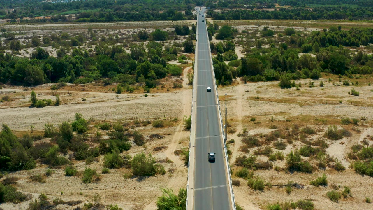 Mid-clip view in San Jose Del Cabo, Mexico showing two cars spaced along the centered road, dry fields and brush beside it, and no visible people.