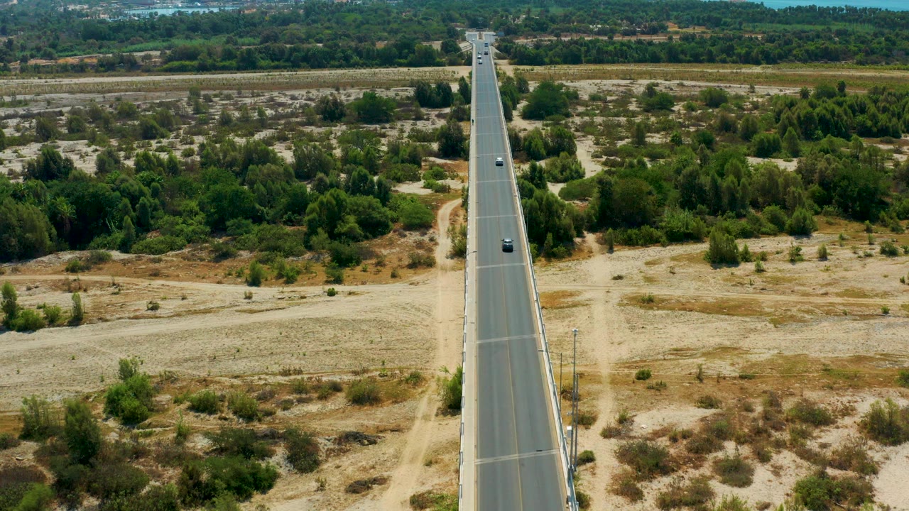 Aerial frame from San Jose Del Cabo, Mexico, showing a straight asphalt road centered through dry scrubland, scattered green trees, and no visible people.