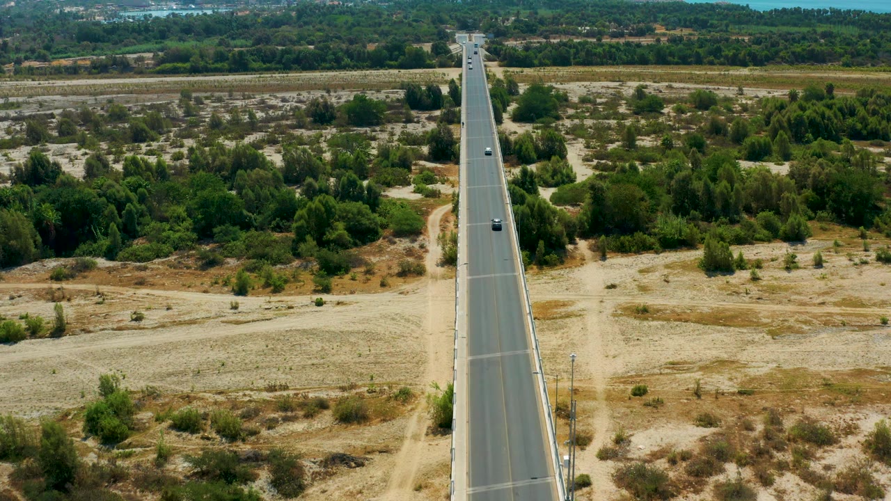 San Jose Del Cabo aerial frame with a few cars on a long straight road, open sandy ground in the foreground, green vegetation behind it, and no visible people.