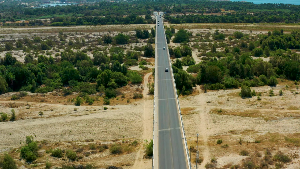Aerial frame from San Jose Del Cabo, Mexico, showing the road slightly left of center, dry shoulder areas, a dirt track, dense green trees, and no visible people.