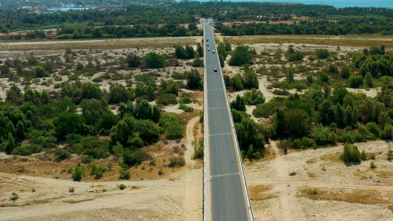 Aerial frame from San Jose Del Cabo, Mexico showing a straight bridge road centered through trees and sandy terrain, with small cars in the distance and no visible people.