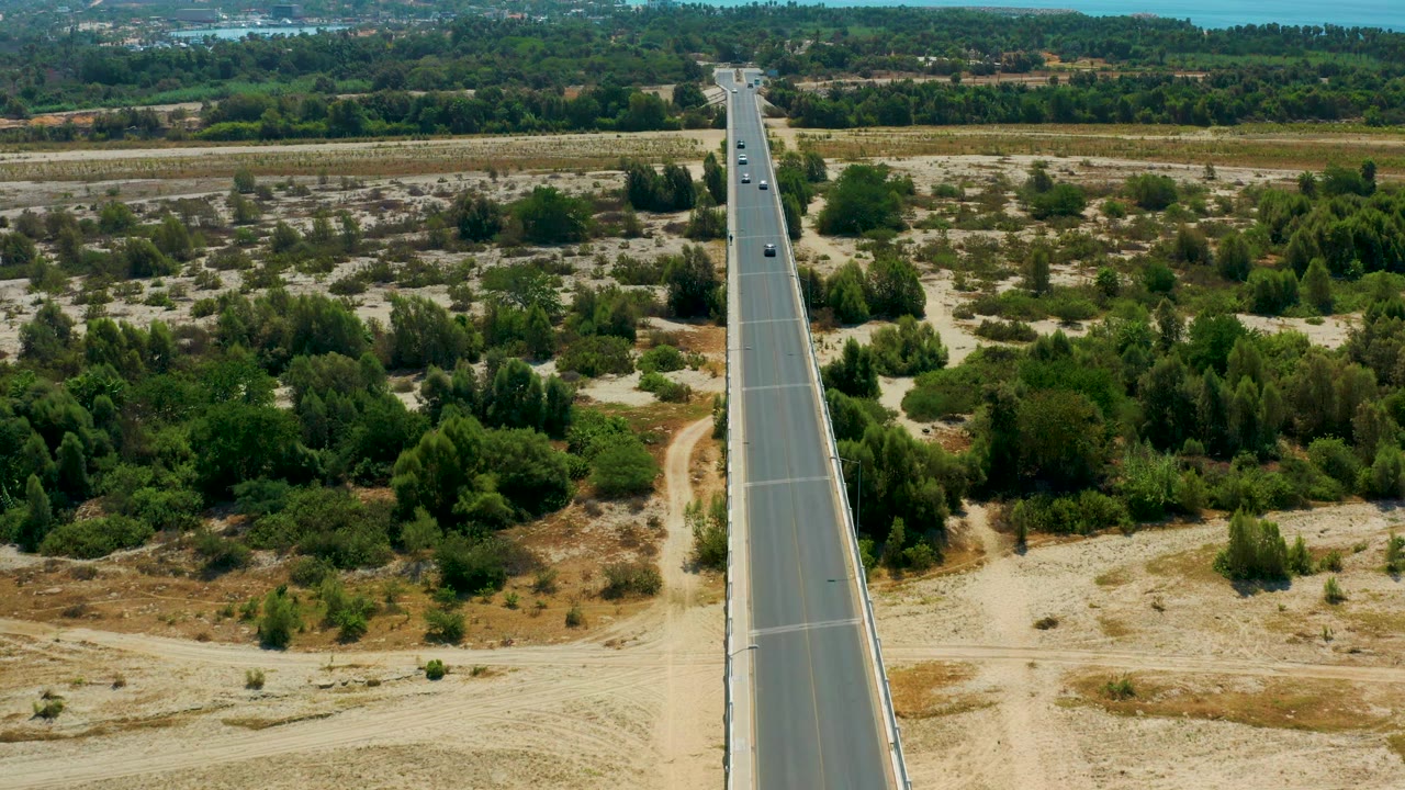 Aerial view in San Jose Del Cabo, Mexico with the bridge slightly nearer in frame, cars spaced along the roadway, green shrubs on both sides, and no visible people.