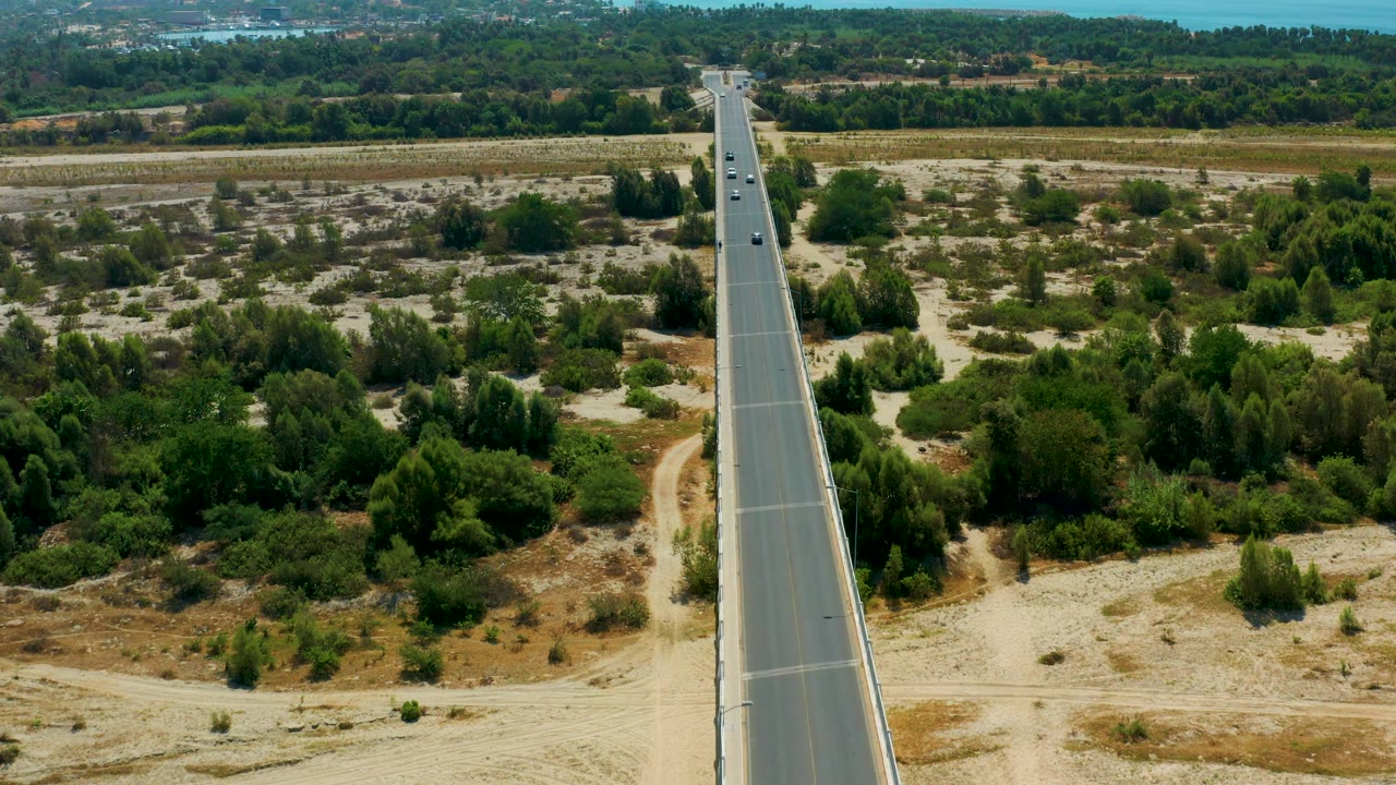 San Jose Del Cabo aerial frame with a long gray bridge dividing scrubby green vegetation and pale ground, several cars visible on the lanes, and no visible people.
