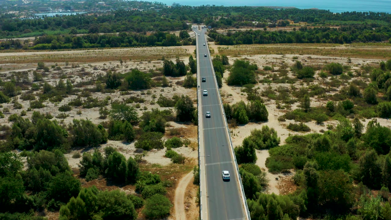 High aerial frame in San Jose del Cabo showing a straight highway centered through sandy scrubland, scattered cars, green tree bands, distant blue water, and no visible people.