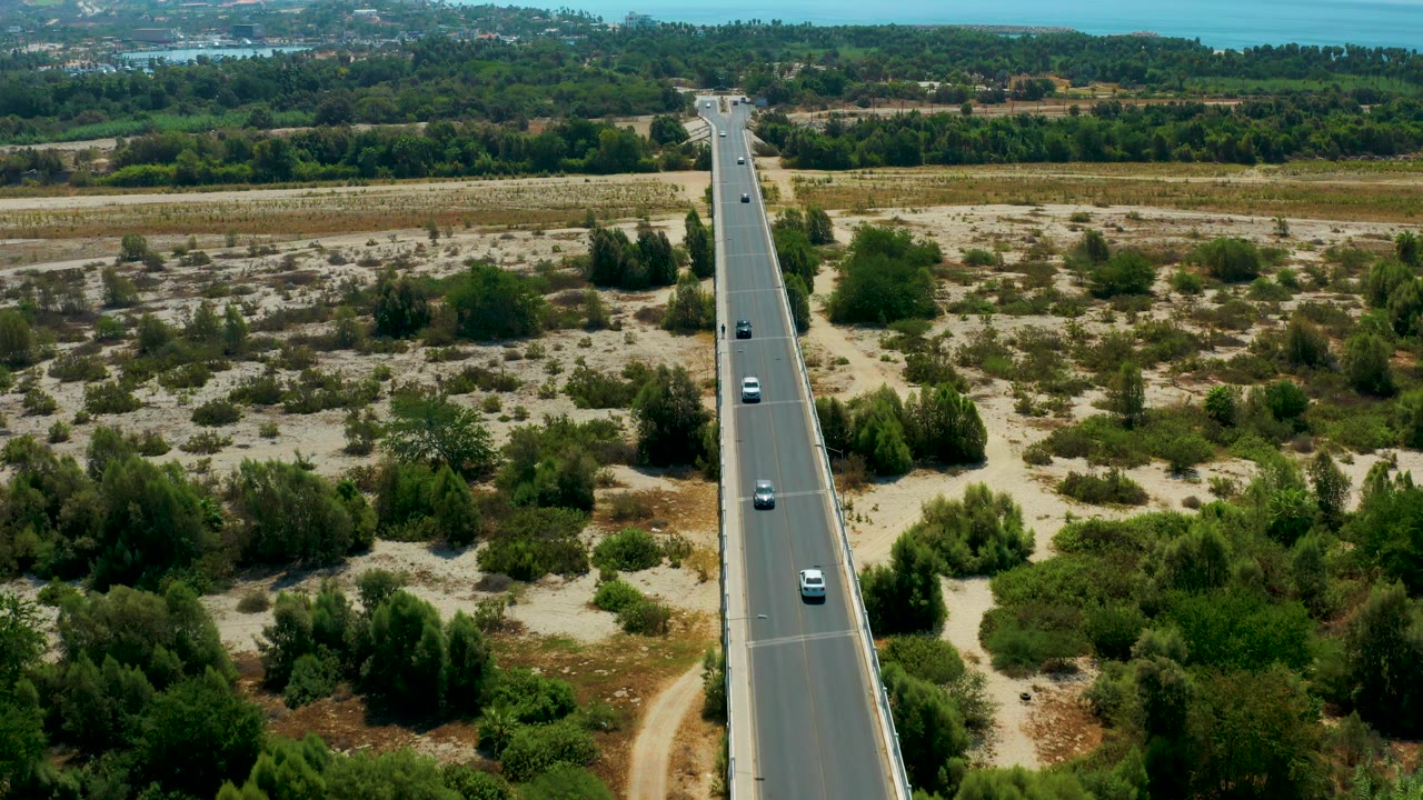Aerial view over San Jose del Cabo with cars on a straight highway, sandy roadside scrub on both sides, dense green trees beyond, distant water, and no visible people.