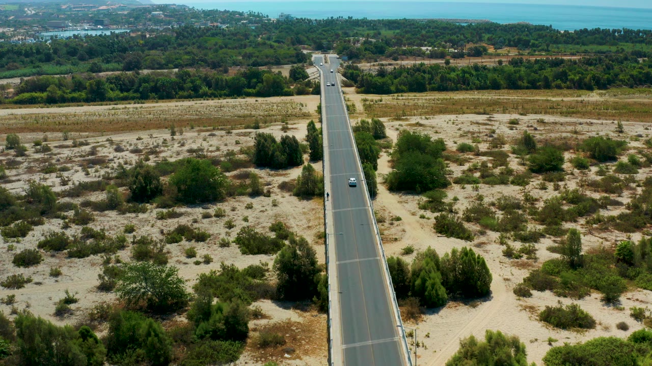 Elevated frame from San Jose Del Cabo, Mexico, showing a straight paved road through sandy scrubland with green trees and distant blue water, no visible people.