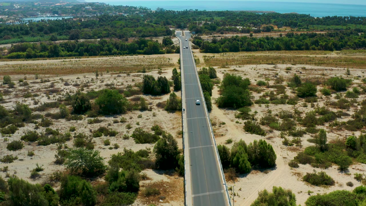 A centered aerial view in San Jose Del Cabo, Mexico, with the paved road dividing dry brush, low green trees, and a distant band of blue water, no visible people.