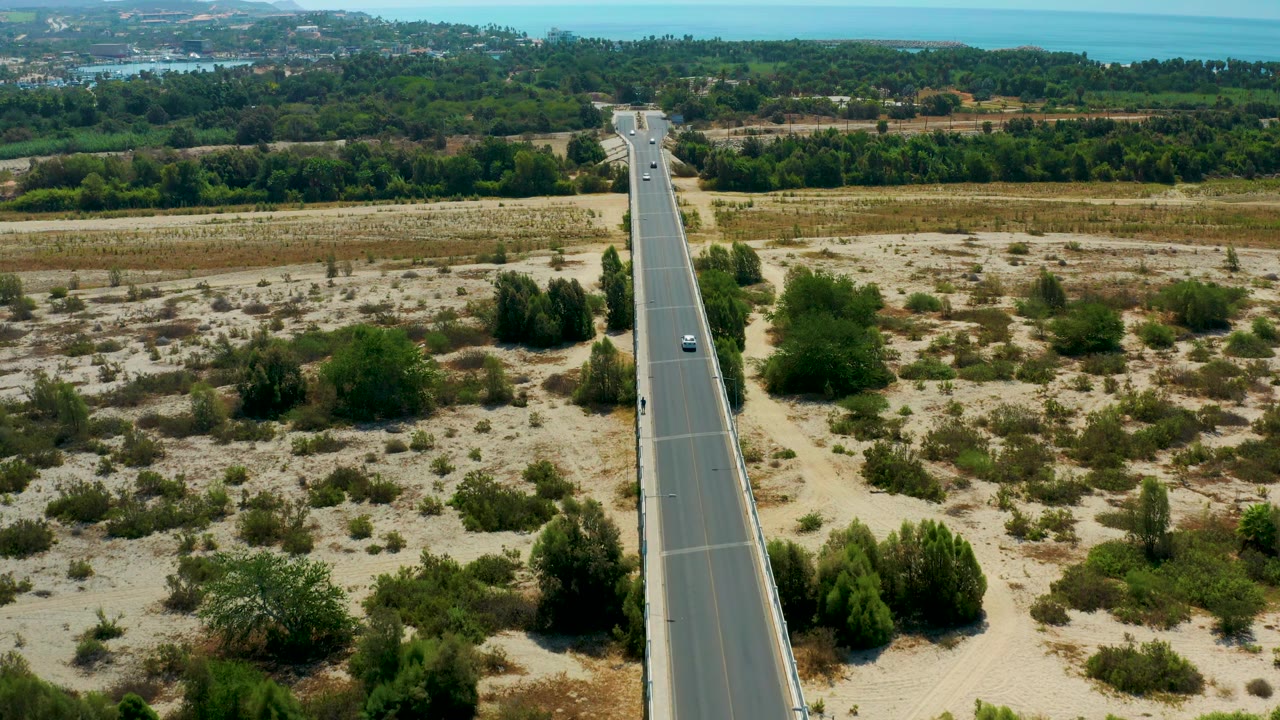 San Jose Del Cabo, Mexico, appears as a long straight road with small cars, pale sandy terrain, scattered bushes, dense trees beyond, and no visible people.