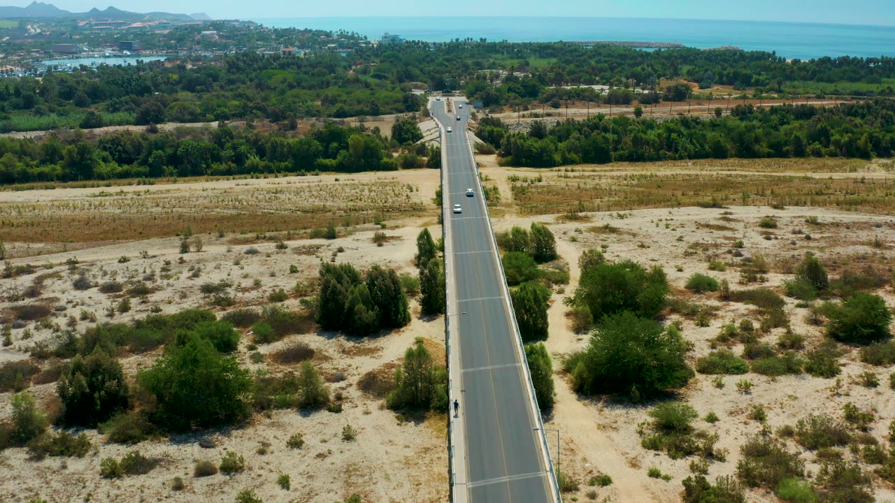 Mid-clip aerial frame over San Jose Del Cabo, Mexico, with the paved road held near the center, pale sand and scrub in the foreground, green trees in back, distant water, and no visible people.