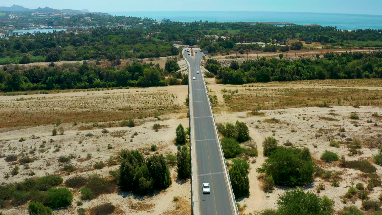 A high aerial view in San Jose Del Cabo, Mexico shows small cars on a paved road bordered by pale scrubland, tree clusters, and ocean on the horizon, with no visible people.