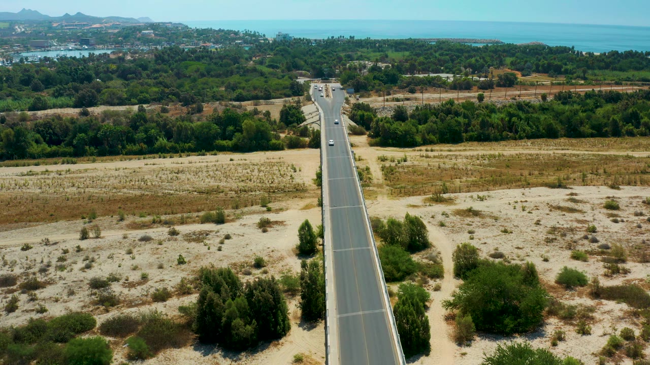 The aerial composition in San Jose Del Cabo, Mexico keeps the road near center as sparse cars pass through scrub, green vegetation, distant development, hills, and blue sea, with no visible people.