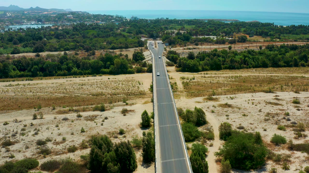A wider moment in San Jose Del Cabo, Mexico shows the road cutting up from the lower frame through tan desert ground toward dense green trees and blue water, with no visible people.