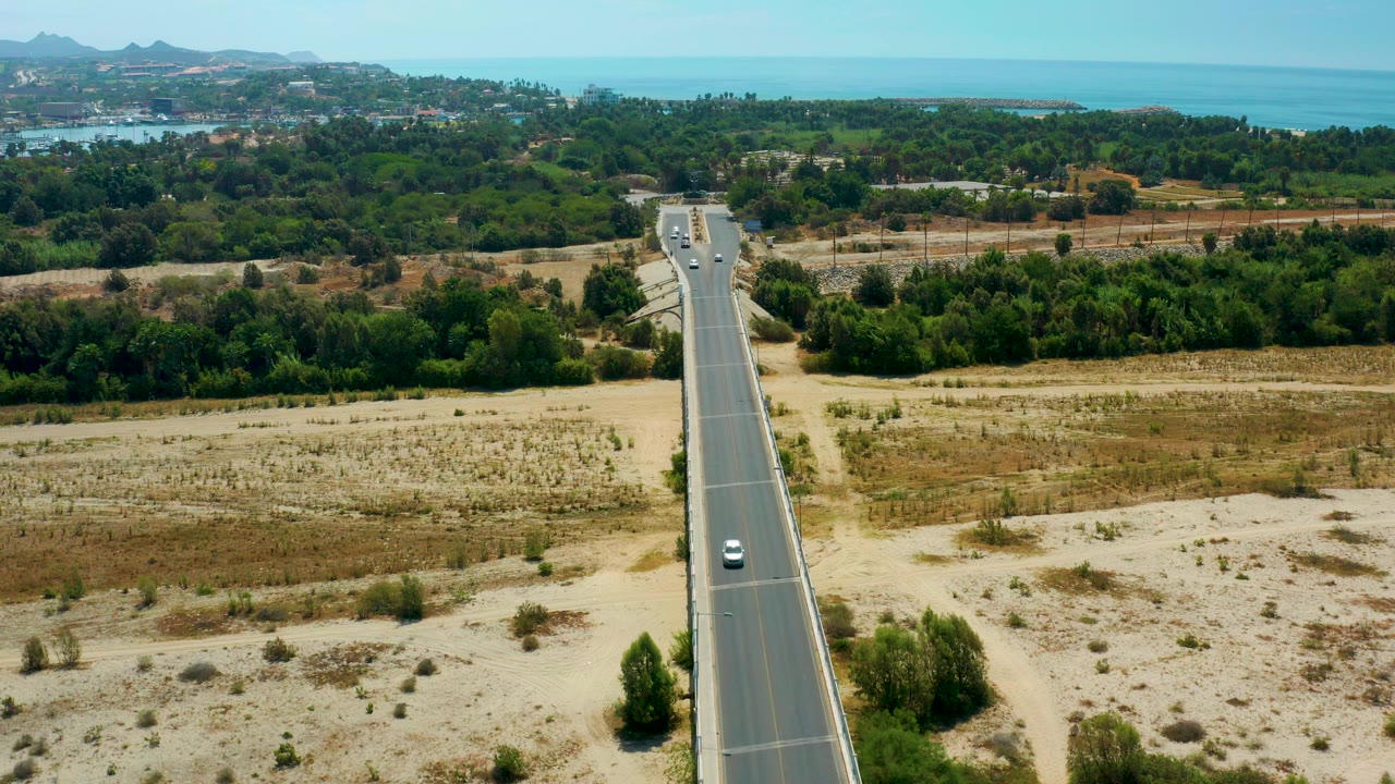 Aerial frame from San Jose Del Cabo showing a straight road centered through dry tan fields and green trees, with blue ocean and distant hills in the background and no visible people.