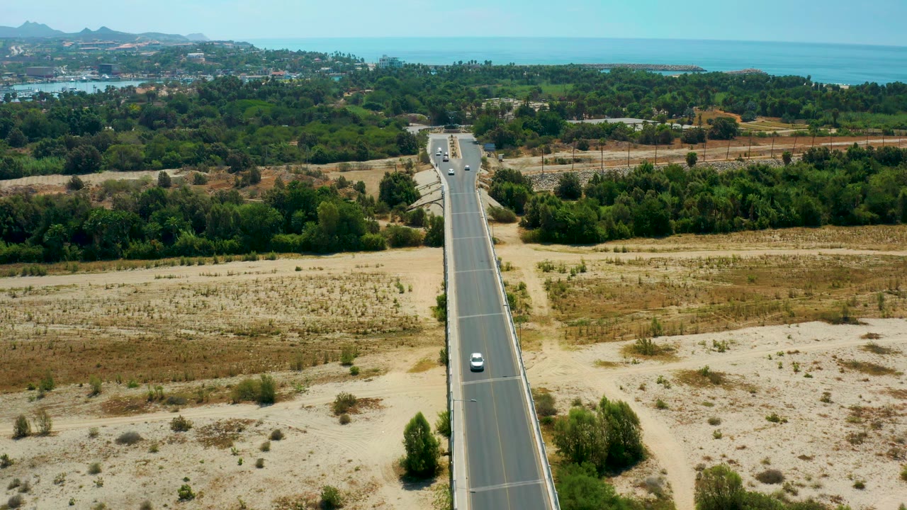 Centered aerial road view with sparse cars, dry grassland, trees, and ocean beyond.
