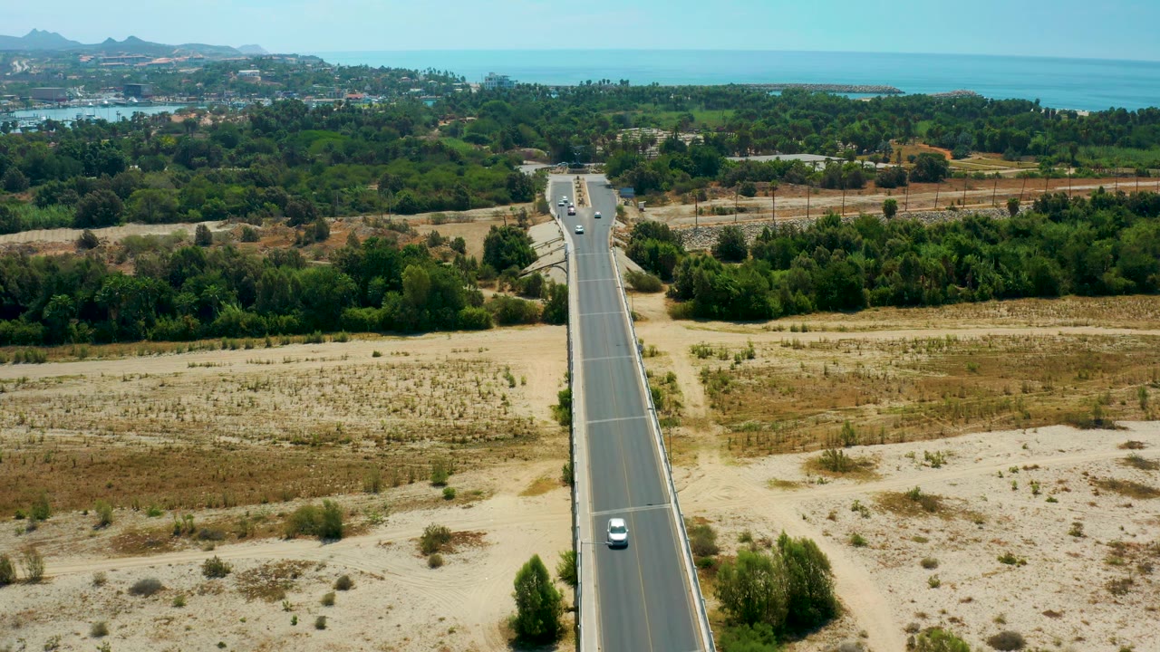 San Jose Del Cabo aerial frame with a small white car lower on the road, pale dry terrain in the foreground, green trees across the midground, and blue ocean behind, with no visible people.