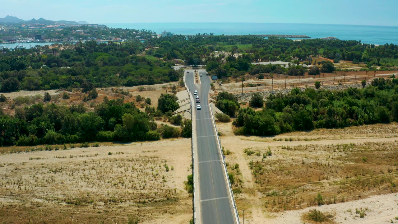Aerial composition over San Jose Del Cabo, Mexico, with the road rising from the bottom edge toward several vehicles, green tree cover, distant water, hills, and no visible people.