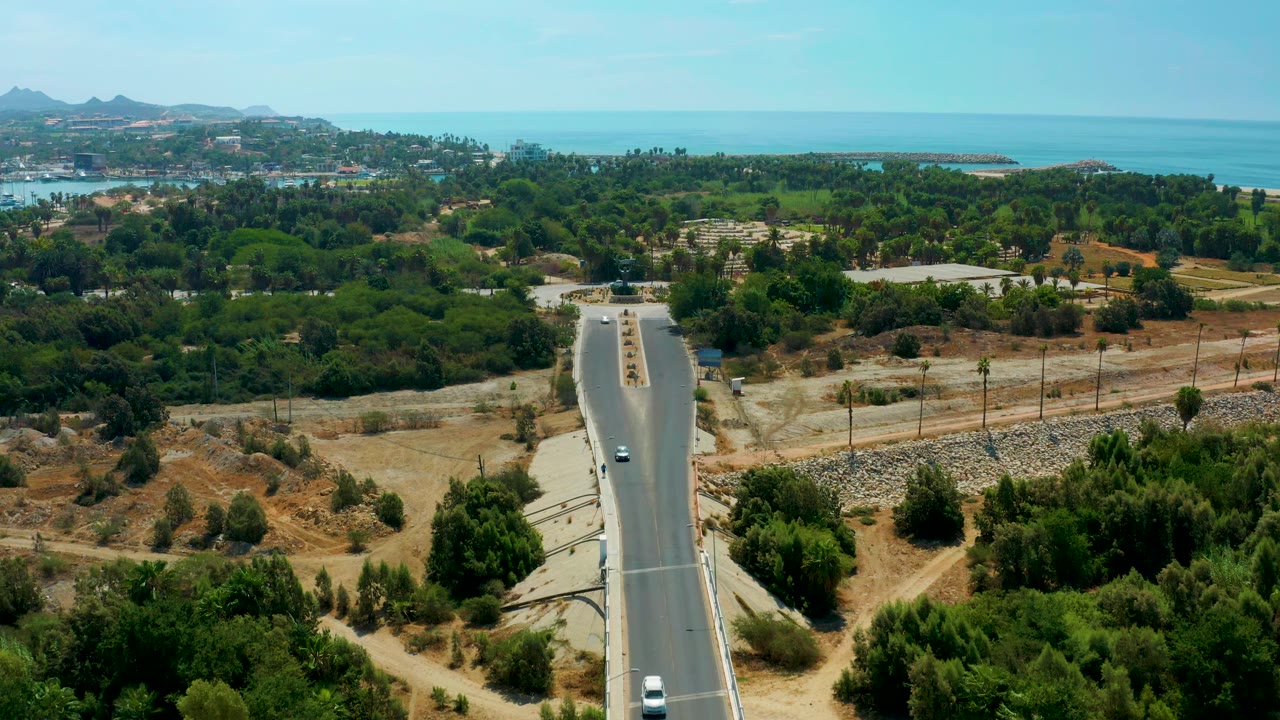 At 0:01, the road remains centered below the drone-like viewpoint, with a few vehicles, dense tree canopy, pale dry lots, and the ocean horizon beyond San Jose Del Cabo, with no visible people.