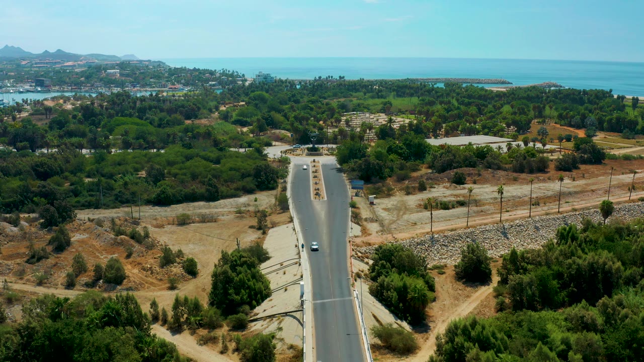 At 0:02, the elevated composition shows vehicles spaced along the divided road, scrubby tan ground beside it, green trees across the middle of frame, and blue water on the horizon, with no visible people.
