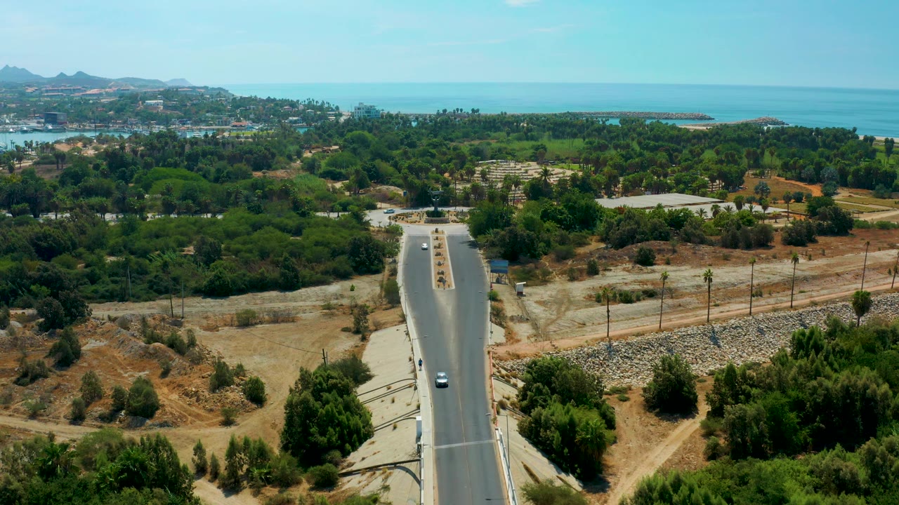 At 0:03, the roadway sits slightly lower in frame with small cars moving through a green corridor, dry open ground to the side, and the San Jose Del Cabo coastline in the distance, with no visible people.