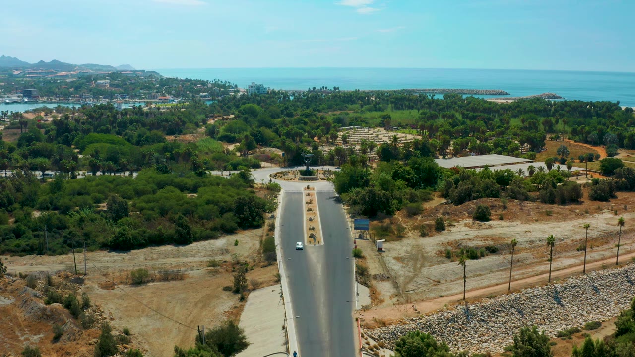 Aerial view in San Jose Del Cabo, Mexico, showing a paved road bordered by dry tan ground and palms, with dense trees, low development, hills, and turquoise-blue water in the background, with no visible people.