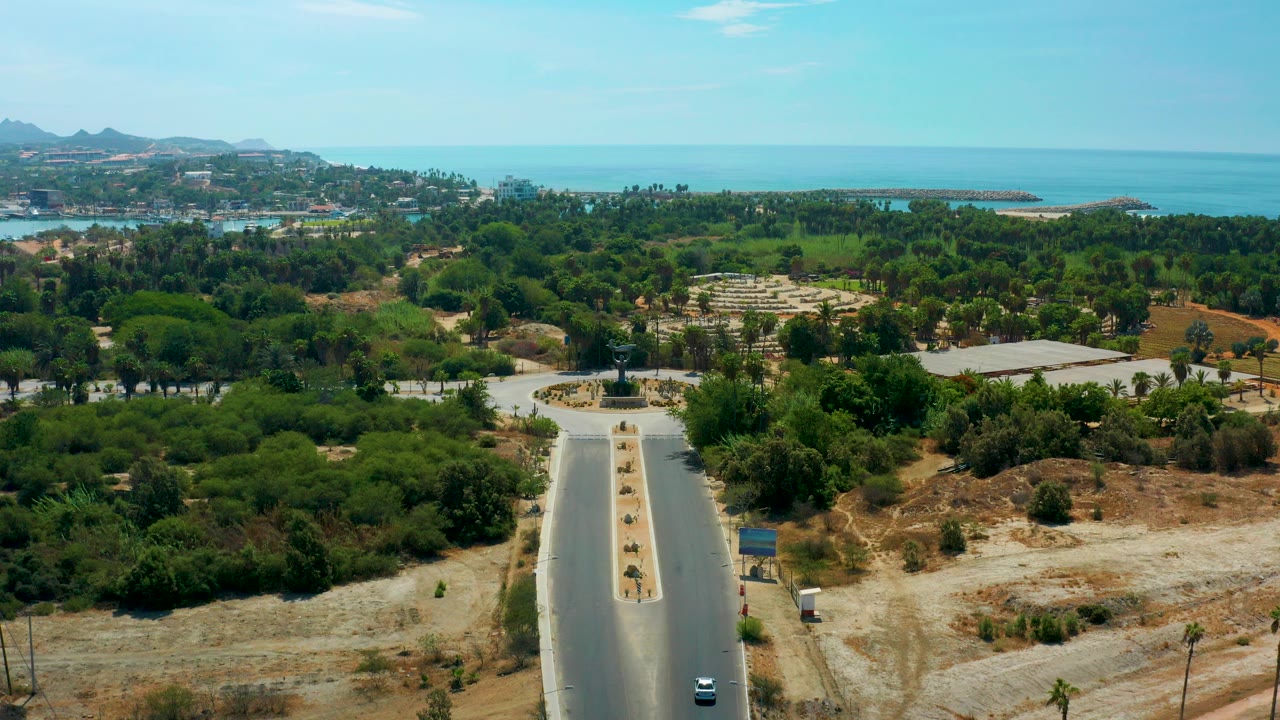 Opening aerial frame in San Jose Del Cabo, Mexico, with a divided road in the foreground, green trees across the midground, blue ocean on the horizon, and no visible people.