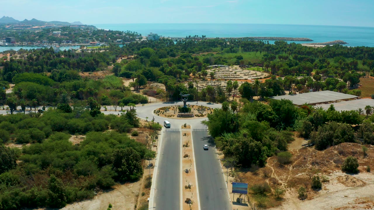 Wide aerial frame over San Jose Del Cabo showing a divided road, a circular roundabout with a central sculpture, dense trees, distant hills, and blue sea, with no visible people.