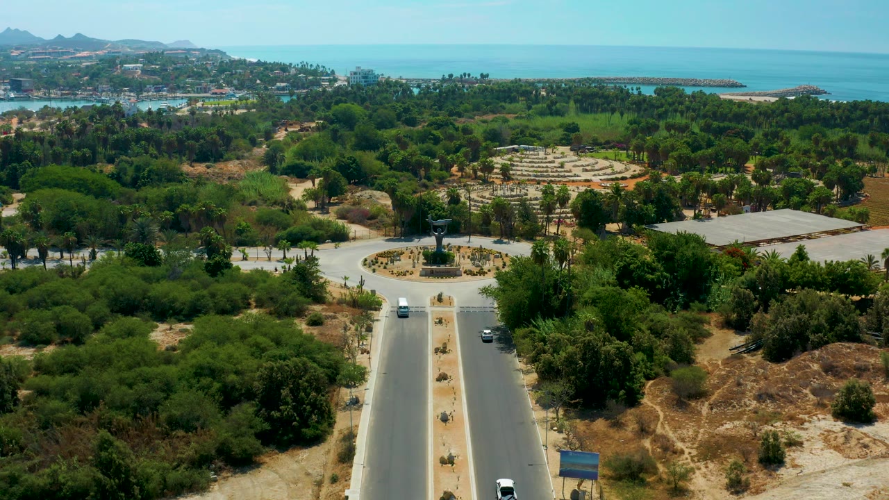 High drone-style frame over San Jose Del Cabo with the divided road centered below, the sculpture roundabout beyond it, dense green trees, dry ground, hills, and turquoise sea, with no visible people.