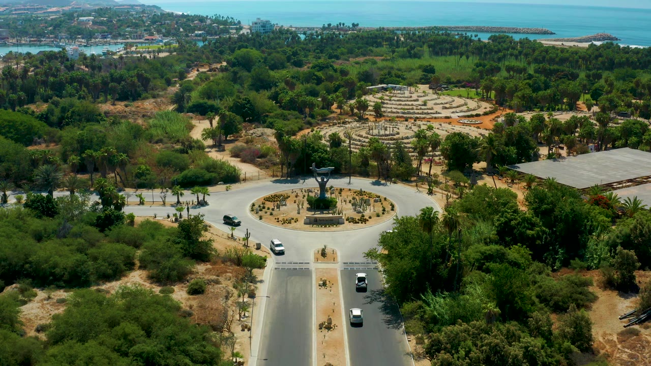 High view of cars circulating around a landscaped roundabout near trees and distant coastline.