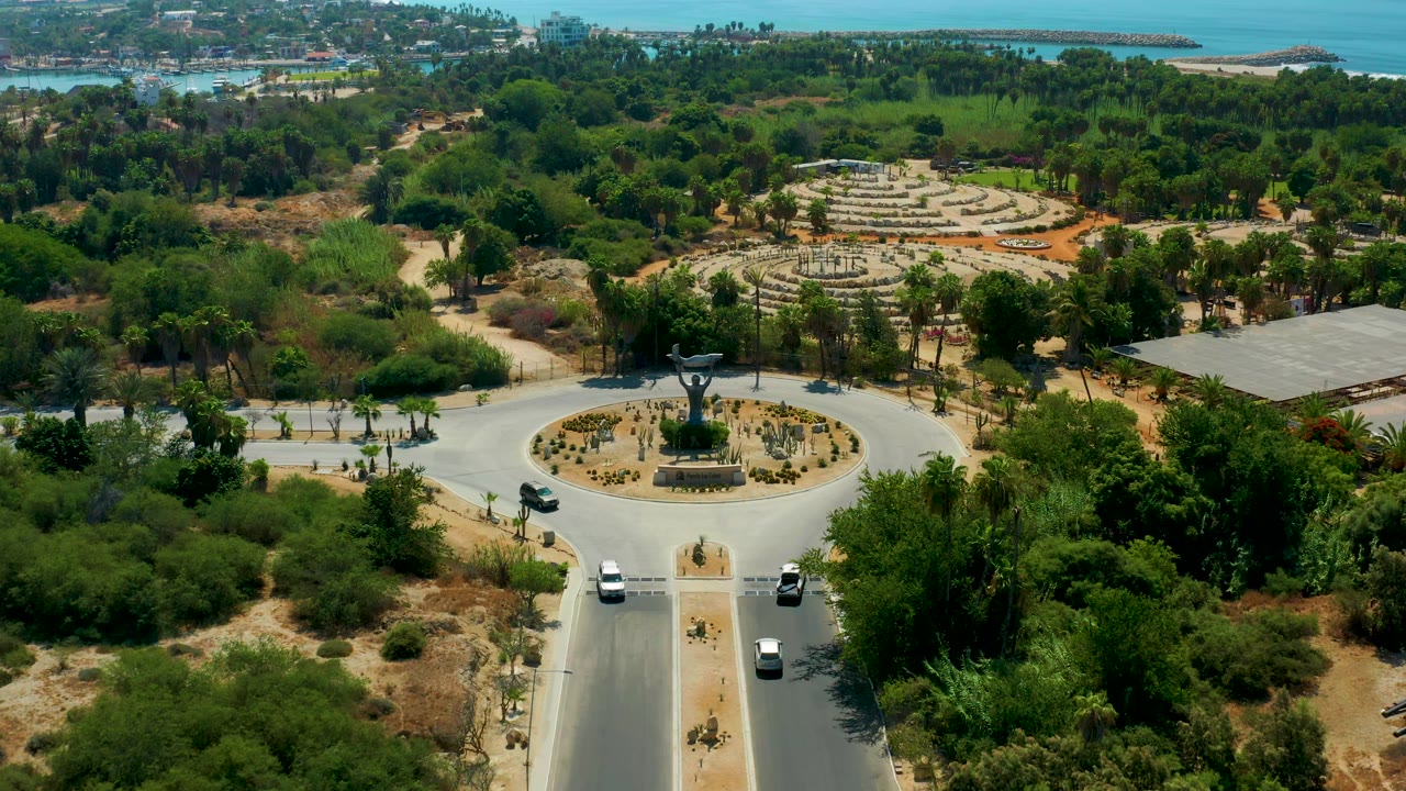 The frame looks down over the San Jose Del Cabo traffic circle, with several cars on the road, palms around sandy medians, dense greenery to the sides, buildings near the water, and no visible people.