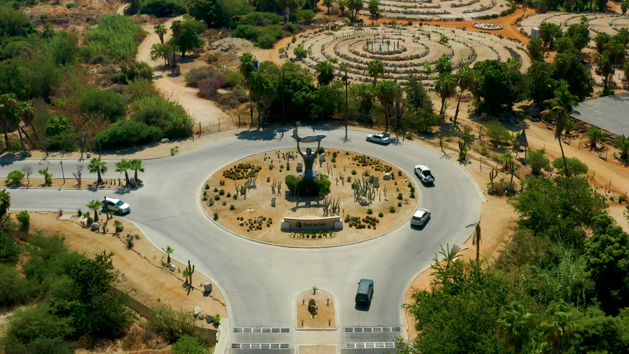 High aerial view in San Jose del Cabo, Mexico, showing several cars around a circular roundabout with a central monument, desert planting, trees, and no visible people.