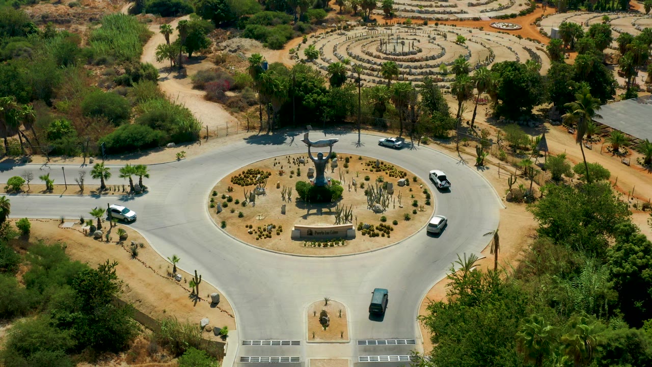 Aerial frame of the San Jose del Cabo traffic circle with white and dark cars on the road, a centered monument, planted island, and no visible people.