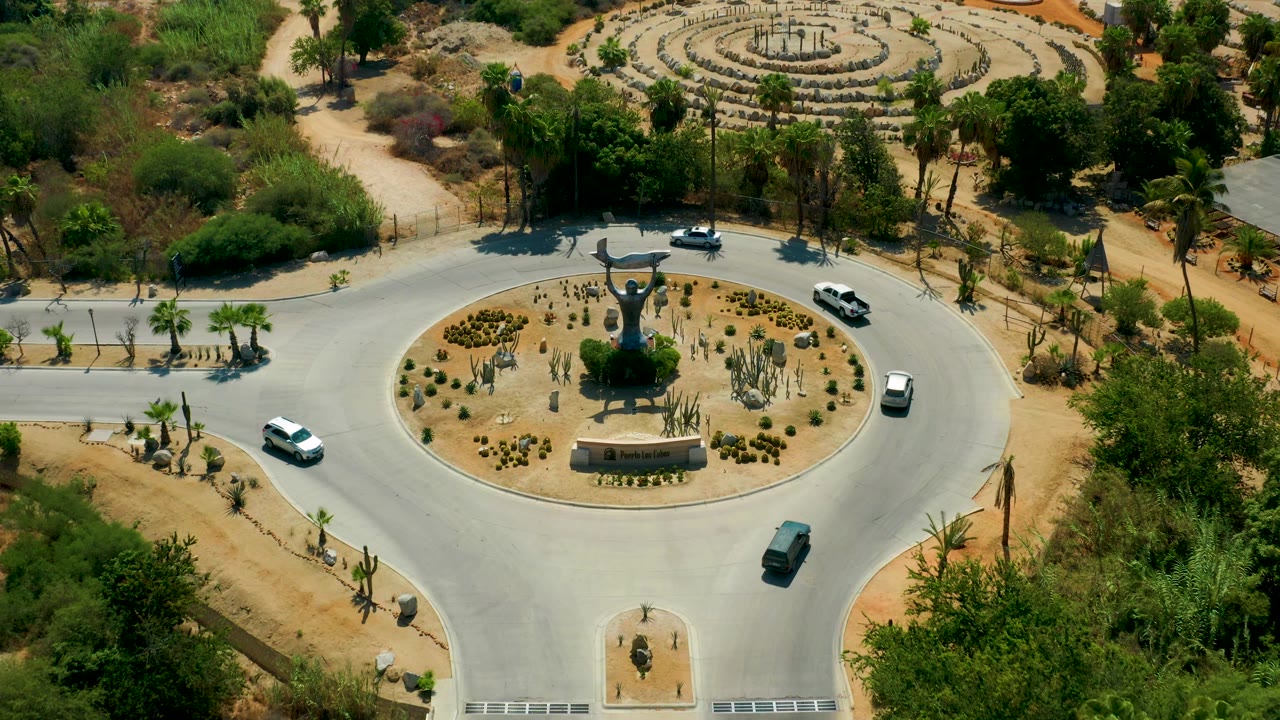Aerial view over the San Jose del Cabo roundabout as vehicles occupy different sides of the circle, with a central sculpture, cacti, shrubs, and no visible people.