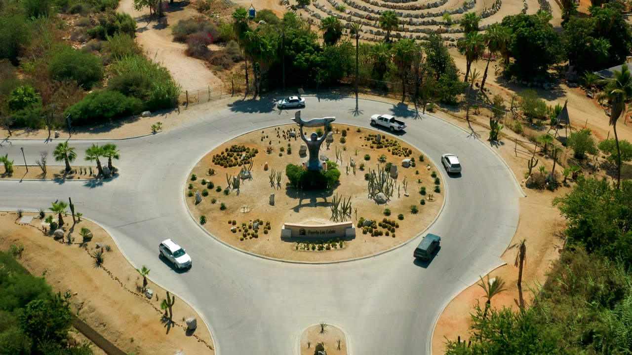 A landscaped roundabout with several cars, cacti, palms, and a central statue.