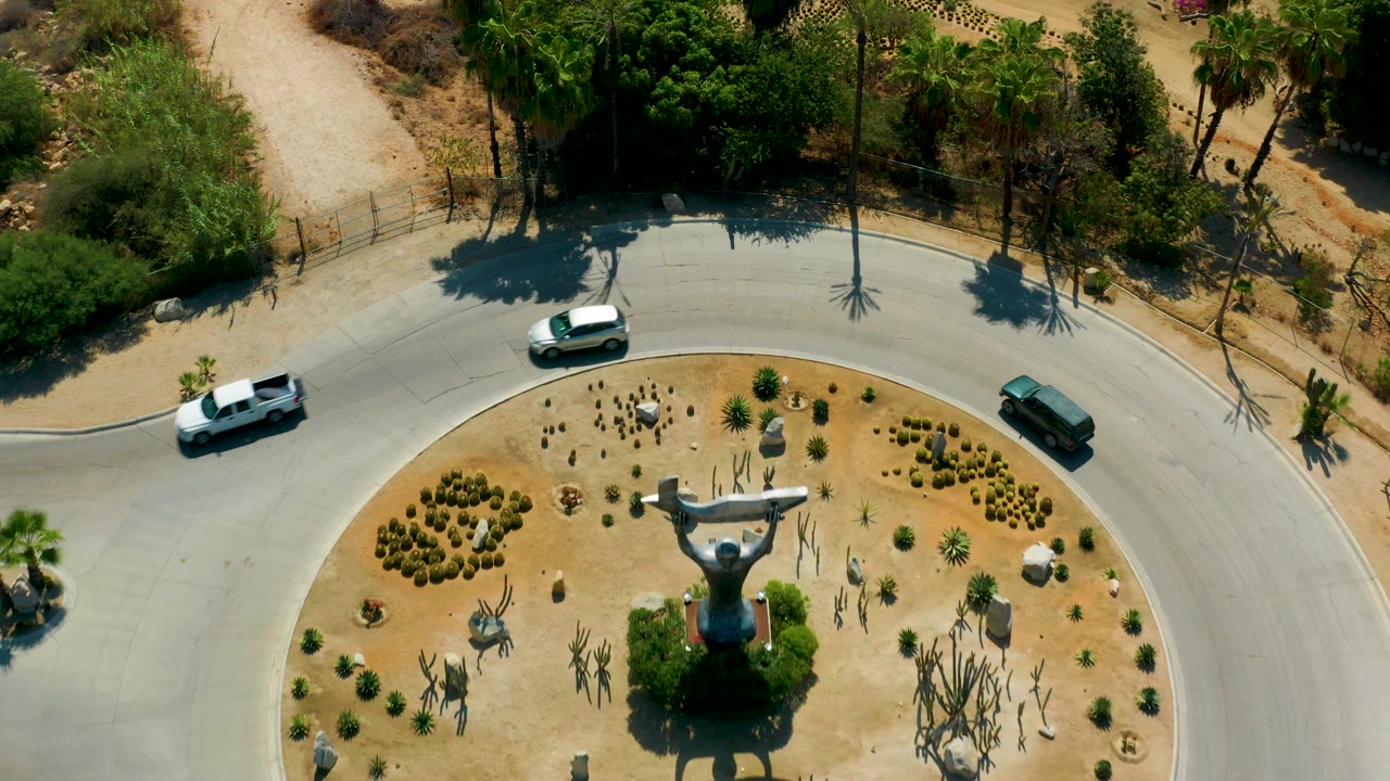 Overhead frame in San Jose Del Cabo, Mexico showing a curved paved road, dry planted circle, central sculpture, trucks, cars, palms, and no visible people.