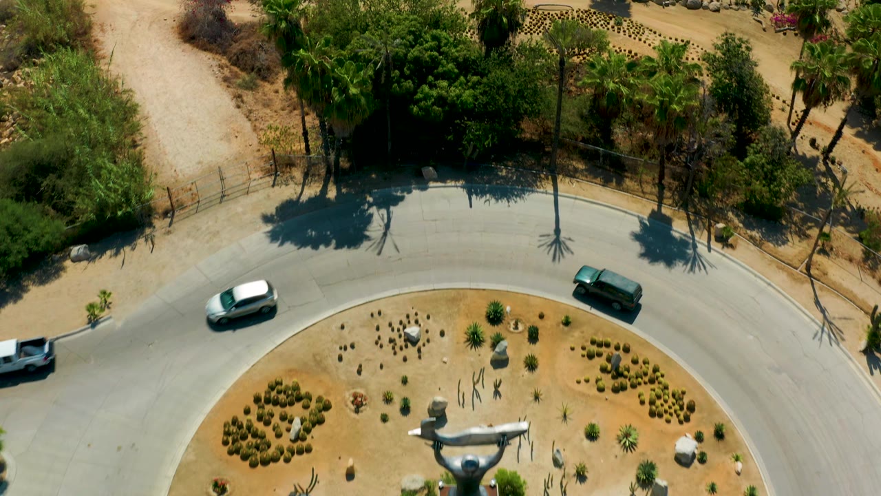 Top-down frame in San Jose Del Cabo, Mexico showing the road, vehicles, planted circle, partial central sculpture, rock terraces, desert paths, and no visible people.