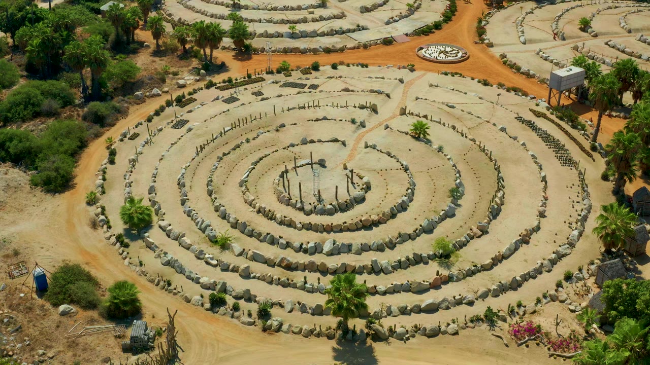Aerial frame over a circular stone-lined forum in San Jose Del Cabo, Mexico, with palms, bushes, dirt paths, and no visible people.