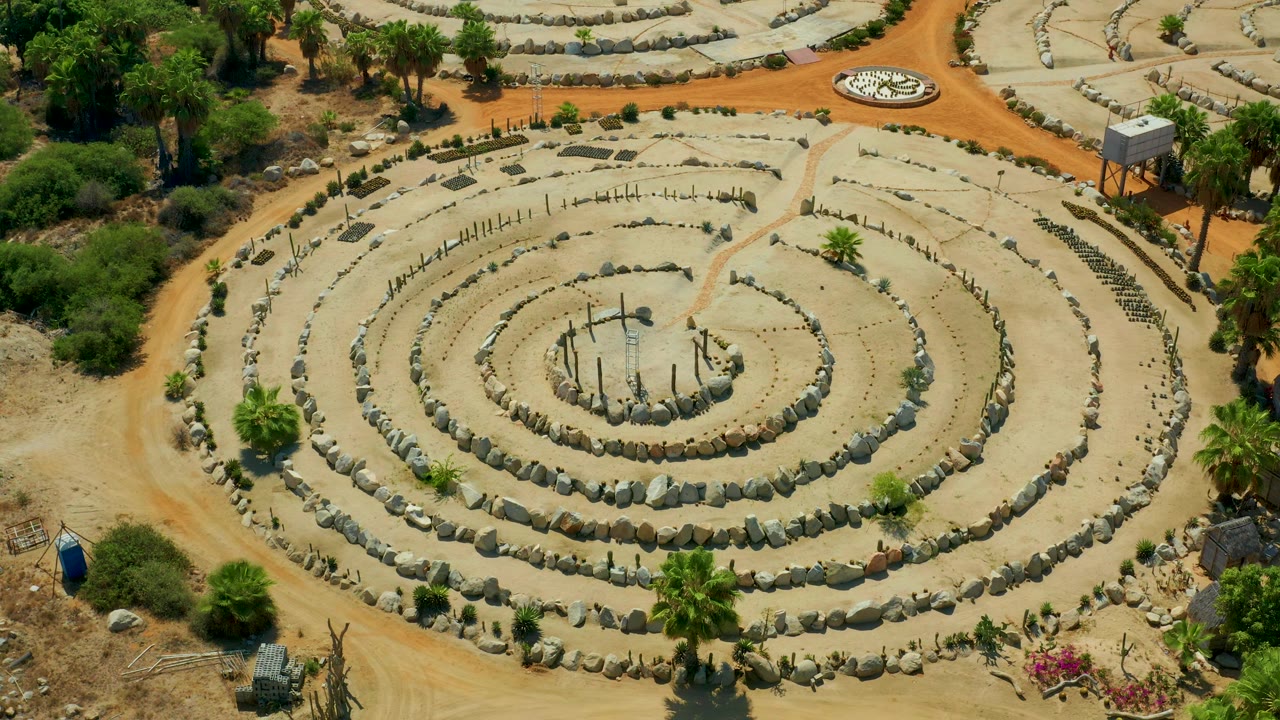 Drone-style overhead frame of the San Jose Del Cabo circular garden design, with reddish dirt paths crossing near the top, low shrubs, palms, and no visible people.
