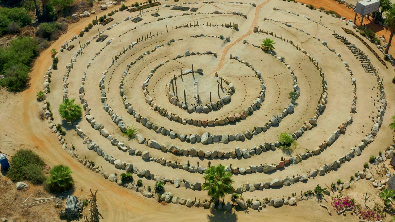 High overhead frame in San Jose Del Cabo, Mexico showing the full sandy stone-ring design, sparse green landscaping, a reddish track along the side, and no visible people.
